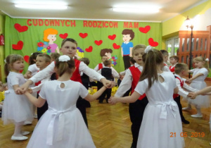 Dziewczynki w białych sukienkach oraz chłopcy w czerwono-czarnych kamizelkach i czarnych spodniach tańczą w parach.